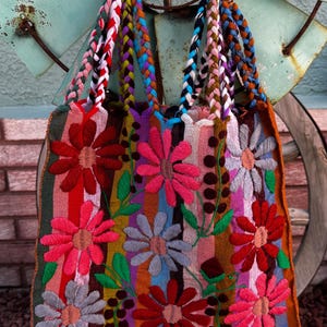 May include: A colorful tote bag with embroidered flowers in shades of red, pink, and gray. The bag features vertical stripes in various colors and braided handles in multiple hues. The background includes a brick wall and a weathered metal windmill.