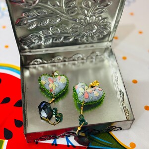 May include: Heart-shaped earrings with a floral pattern in a silver metal box. The earrings have a light blue background with colorful flower designs and green trim. Each earring has a dangling dark green crystal and gold-colored accents. The box lid is decorated with embossed floral patterns.