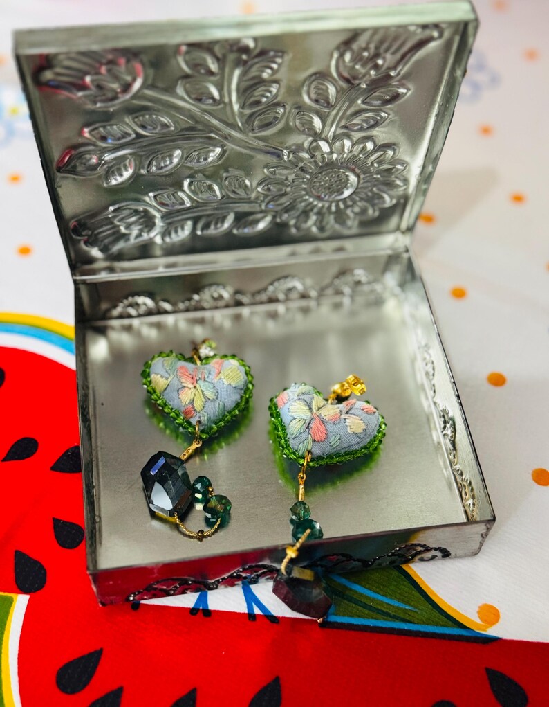 May include: Heart-shaped earrings with a floral pattern in a silver metal box. The earrings have a light blue background with colorful flower designs and green trim. Each earring has a dangling dark green crystal and gold-colored accents. The box lid is decorated with embossed floral patterns.