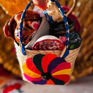 May include: A small woven basket with a blue handle and a colorful flower design on the front. The basket is filled with miniature ceramic items, including a small pitcher and bowls, showcasing a vibrant display of Mexican folk art.