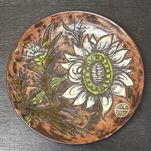 Peut inclure: Une assiette en céramique brune avec un motif floral blanc et vert. L'assiette a un bord brun et une étiquette blanche qui dit "Ruscha Handgemacht".