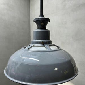 Op de afbeelding: Een glanzend grijze hanglamp met een brede, koepelvormige metalen kap en een witte binnendiffuser. De lamp hangt aan een zwarte stang en is voorzien van een donkergrijze montagekap met een klein messing schroefdetail aan de bovenzijde.