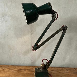 May include: A dark green metal articulated desk lamp with a bell-shaped shade and a rectangular base. The lamp features a multi-jointed arm connected by visible bolts and is powered by a red fabric-covered electrical cord with a toggle switch.