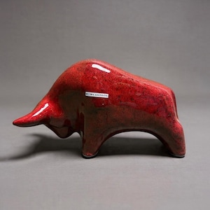 May include: A glossy red ceramic bull figurine in a charging stance with a lowered head. The surface features a mottled, textured finish with darker red and black undertones. A small white rectangular label with the text OROKERAMIK is visible.