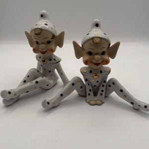 May include: Two ceramic elf figurines. The elves are white with black and gold polka dots. They have pointed hats, large ears, and rosy cheeks. The figurines are in a seated position with their legs spread.