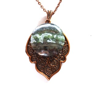 May include: A copper-colored pendant necklace featuring a large, oval-shaped stone with green and gray patterns. The stone is set in a decorative, openwork copper frame and hangs from a matching chain. The pendant has an ornate, vintage-inspired design.