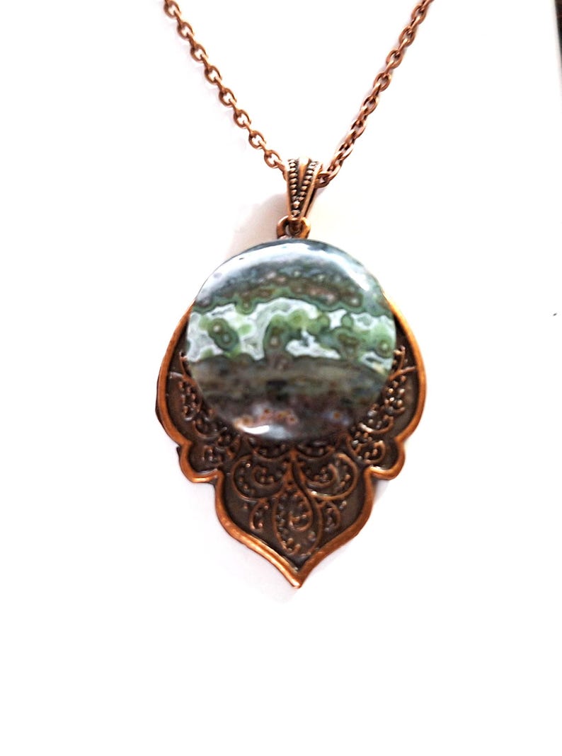 May include: A copper pendant necklace with an oval stone displaying green and grey patterns. The stone is set in an ornate, openwork copper frame, suspended from a matching chain. The pendant has a vintage-inspired design, suitable for everyday wear or special occasions.