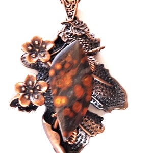 May include: A copper-toned pendant necklace featuring a central, speckled, brown and orange stone. The pendant is adorned with floral and leaf-like details, and hangs from a delicate chain. The design has an antique, handcrafted aesthetic.