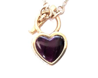 May include: A gold-toned necklace featuring a heart-shaped pendant with a deep purple center. The pendant is framed in gold and attached to a delicate chain. The necklace has a toggle clasp.