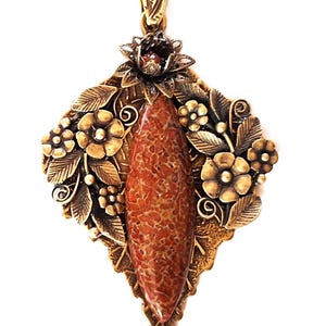 Puede incluir: Un collar con colgante de estilo vintage con un diseño detallado y ornamentado. El colgante presenta una piedra central, de color marrón rojizo y forma ovalada, rodeada de metal dorado con motivos florales y de hojas. El collar tiene una cadena delicada.