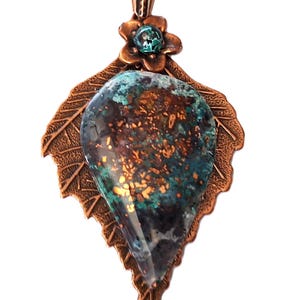 May include: Copper pendant necklace featuring a large, teardrop-shaped stone with teal, brown, and gold tones. The stone is set in a copper leaf-shaped frame with a flower accent and a delicate chain.