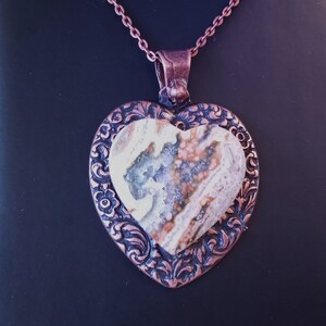 Puede incluir: Un colgante de corazón de cobre con un intrincado diseño floral. El colgante presenta una piedra de ágata natural con remolinos marrones, blancos y grises.