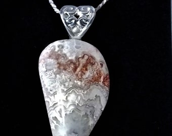 May include: A silver chain necklace with a heart-shaped pendant featuring an intricate design. The pendant is attached to a large, teardrop-shaped stone with a white and brown agate pattern.