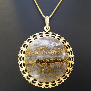 Puede incluir: Collar con colgante dorado con una piedra redonda con tonos grises, marrones y amarillos. La piedra está engastada en un marco calado dorado y cuelga de una fina cadena dorada. El diámetro de la piedra es de aproximadamente 4 cm.