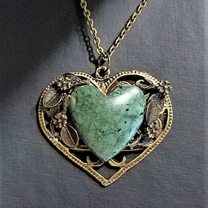 Puede incluir: Un collar con un colgante en forma de corazón dorado adornado con una piedra verde en forma de corazón en el centro. El colgante está decorado con intrincados detalles florales.