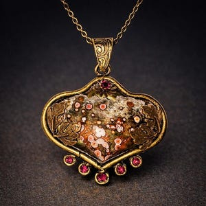 May include: A brass-toned pendant necklace featuring a central heart-shaped stone with brown, white, and red circular patterns. The metal frame is decorated with leaf motifs and small, round, deep pink gemstones along the bottom edge.