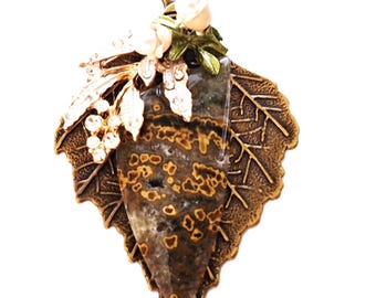 May include: A leaf-shaped pendant necklace. The pendant features a central stone with brown and tan patterns, set within a bronze-coloured leaf. It is adorned with white flowers and clear crystals. The necklace chain is bronze.