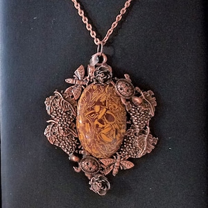 May include: A copper necklace with a pendant featuring a brown and orange oval stone surrounded by copper leaves, grapes, and turtles.