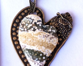 Puede incluir: Un collar con un colgante en forma de corazón y una cadena marrón. El colgante está hecho de dos corazones, uno con una superficie texturizada y el otro con una piedra de color marrón claro, verde y blanco.
