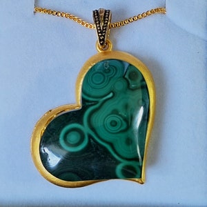 Puede incluir: Un collar con un colgante en forma de corazón de oro y una piedra de malaquita verde. La piedra tiene un patrón de remolino de verde y negro.