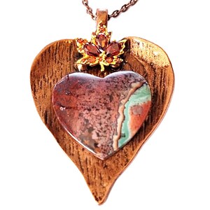 May include: A copper-toned pendant necklace featuring a heart-shaped stone pendant with red, green, and brown hues. The pendant is set within a leaf-shaped frame and adorned with a small, jeweled accent. The necklace chain is also copper-toned.
