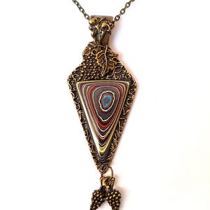 Puede incluir: Un collar con un colgante de oro con una piedra de ágata triangular engastada en un marco detallado y ornamentado. El colgante presenta un diseño de vid y dos racimos de uvas colgantes.