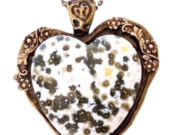 Puede incluir: Collar con colgante en forma de corazón con un centro de piedra moteada en tonos blancos, verdes y amarillos. El colgante está enmarcado por un metal color bronce con detalles florales y cuelga de una cadena a juego. Una joya única.