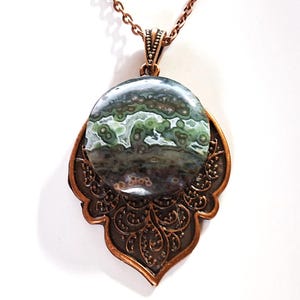 May include: A pendant necklace with a copper-toned chain and setting. The pendant features a large, circular stone with a mottled pattern in green, grey, and brown. The copper setting has an intricate, decorative design.