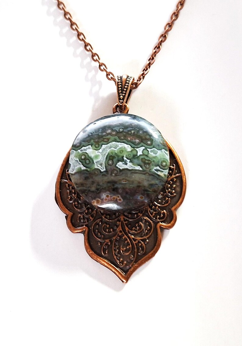 May include: A pendant necklace with a copper-toned chain and setting. The pendant features a large, circular stone with a mottled pattern in green, grey, and brown. The copper setting has an intricate, decorative design.