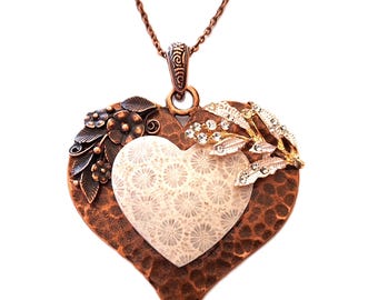 Puede incluir: Un collar con un colgante de corazón de cobre, con un centro blanco de coral y detalles decorativos florales y de hojas. El collar tiene una cadena delicada.