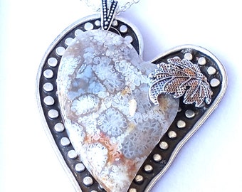 Puede incluir: Collar con colgante en forma de corazón con cadena de plata. El colgante presenta una piedra en forma de corazón con un patrón único, enmarcada por un borde plateado con pequeños acentos redondos. Un detalle de hoja plateada está en el lateral.