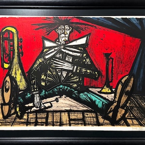 May include: Abstract print of a musician playing a trumpet, set against a red background. The artwork features bold black lines, with accents of yellow, teal, and brown. The print is framed in a black frame.
