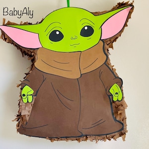K&ouml;nnte beinhalten: Eine gr&uuml;ne und braune Baby Yoda Pi&ntilde;ata mit rosa Ohren. Die Pi&ntilde;ata ist nach der Figur aus der Star Wars-Serie geformt.