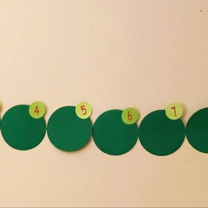 May include: A green caterpillar with red head and yellow eyes, made of paper, with numbers 1 through 12 on each segment. The caterpillar is hanging on a white wall.