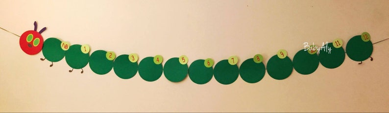 May include: A green caterpillar with red head and yellow eyes, made of paper, with numbers 1 through 12 on each segment. The caterpillar is hanging on a white wall.