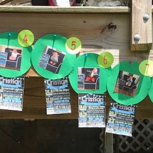 May include: A green and red paper caterpillar banner with numbers 1 through 11 on each segment. The banner is decorated with photos of a baby boy. The banner is hanging on a wooden surface. The text "Cristian" is visible on a sign below the banner.