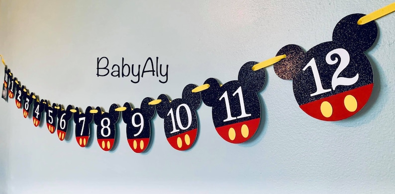 以下が含まれることがあります： 黒と赤のミッキーマウスをテーマにしたバナーで、白い数字が1から12まで並んでいます。バナーは黄色の紐でつなげられています。