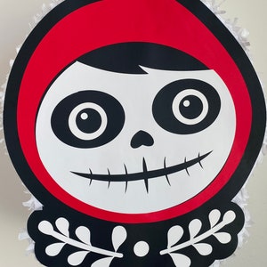 Könnte beinhalten: Eine rote und schwarze Papier-Piñata in Form eines Skeletts, das eine rote Kapuze trägt. Die Piñata hat weiße Augen und ein weißes Lächeln. Die Piñata ist mit weißen Blumenmustern verziert.
