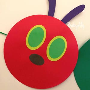 May include: A red felt circle with two green eyes with yellow outlines and a brown nose. The circle is attached to two purple felt antennae. The text "Baby Aly" is visible in the background.
