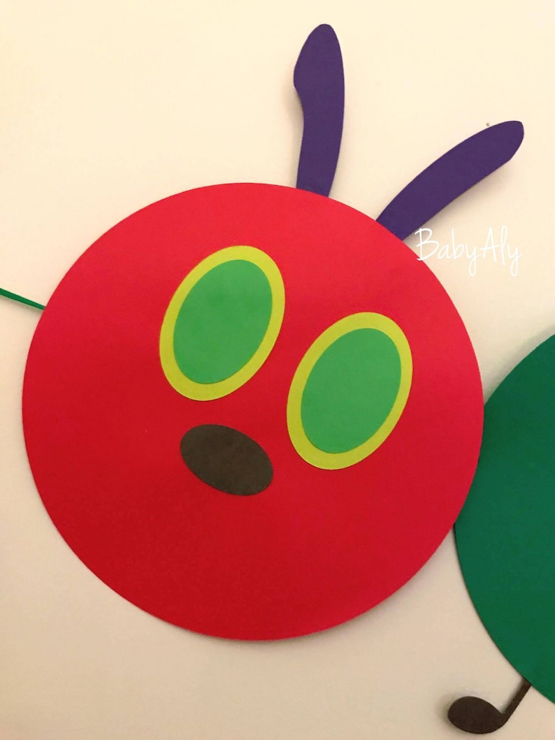 May include: A red felt circle with two green eyes with yellow outlines and a brown nose. The circle is attached to two purple felt antennae. The text "Baby Aly" is visible in the background.