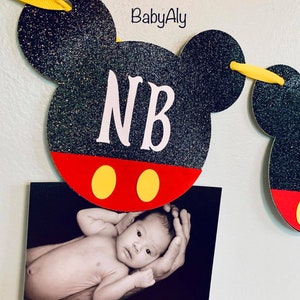 以下が含まれることがあります： 黒と赤のミッキーマウスをテーマにしたバナーで、白い文字で「NB」と書いてあります。バナーには黄色のリボンタイが付いており、赤ちゃんの写真がバナーに付けられています。