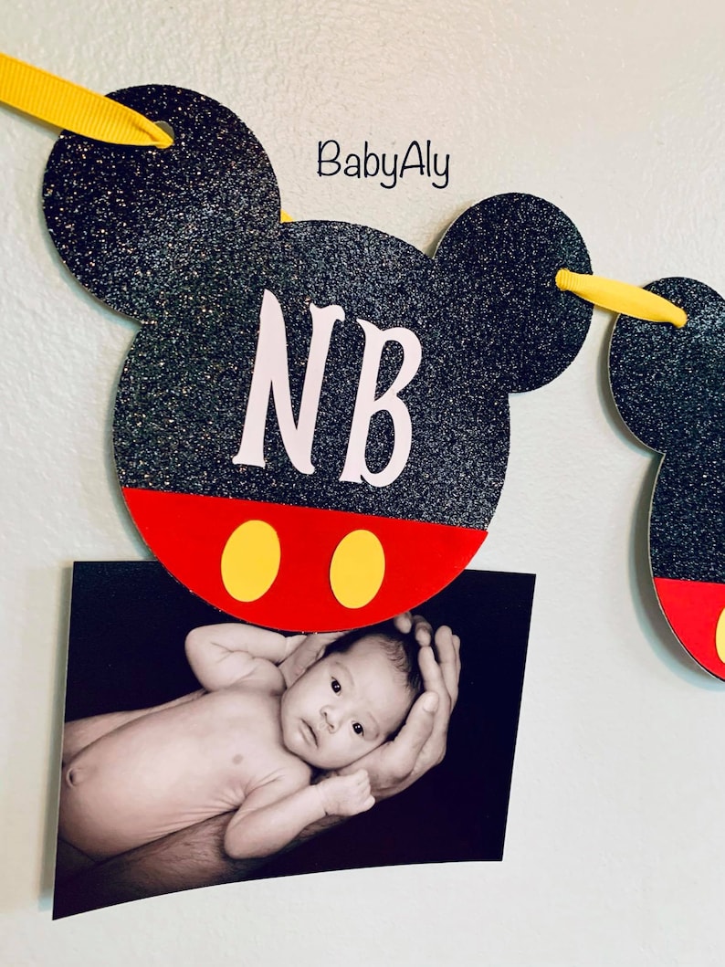 以下が含まれることがあります： 黒と赤のミッキーマウスをテーマにしたバナーで、白い文字で「NB」と書いてあります。バナーには黄色のリボンタイが付いており、赤ちゃんの写真がバナーに付けられています。