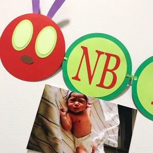 May include: A red, green, and purple paper caterpillar banner with the letters "NB" on one of the segments. A photo of a baby is attached to the banner.