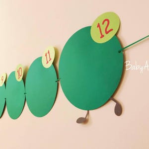 May include: Green paper circles with red numbers 9 through 12, connected by string to form a caterpillar shape. The caterpillar has gray legs and the text "Baby Aly" is written on the wall.
