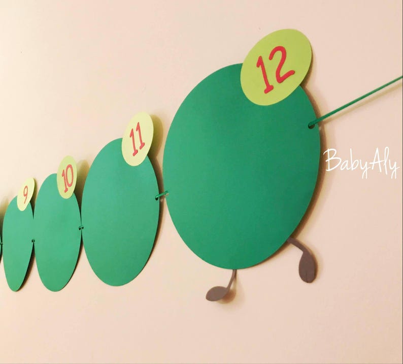 May include: Green paper circles with red numbers 9 through 12, connected by string to form a caterpillar shape. The caterpillar has gray legs and the text "Baby Aly" is written on the wall.