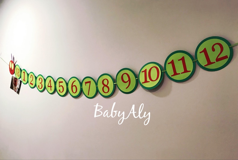 May include: A green and red felt banner with the numbers 1 through 12 in red circles. The banner is shaped like a caterpillar and has a red head with a face. The text "Baby Aly" is written below the banner.