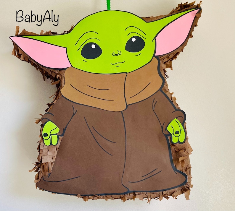 K&ouml;nnte beinhalten: Eine gr&uuml;ne und braune Baby Yoda Pi&ntilde;ata mit rosa Ohren. Die Pi&ntilde;ata ist nach der Figur aus der Star Wars-Serie geformt.