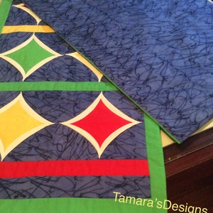 May include: A blue table runner with a diamond pattern in yellow, green, red and white. The runner has a green border and the text "Tamara's Designs" is visible in the bottom right corner.