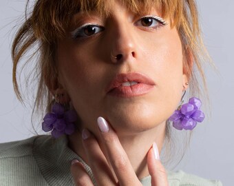 May include: A pair of purple flower-shaped earrings featuring translucent, multi-faceted petals that catch the light. The earrings hang from silver-toned metal hooks and are designed with a layered, clustered petal structure that creates a dimensional look.