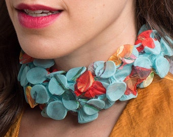 Puede incluir: Primer plano de un collar con diseño floral. El collar presenta círculos de tela azul claro con estampados florales rojos, naranjas y amarillos. El collar se lleva sobre un top amarillo-naranja.